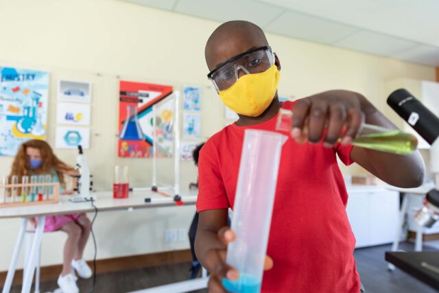 photo - Boy Wearing Face Mask and Protective Glasses Mixing Chemicals in Science Class