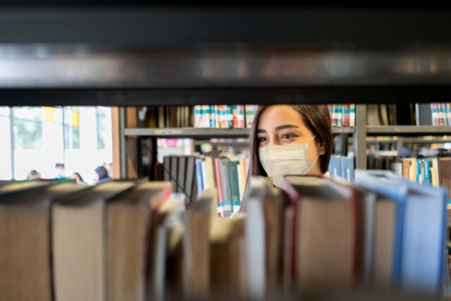 photo - Student Wearing a Facemask at the Library While Looking for a Book